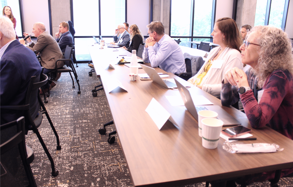 Audience members sit around a conference table in a bright meeting room, listening to a speaker holding a microphone; laptops, notebooks, and coffee cups are visible on the table near large windows.