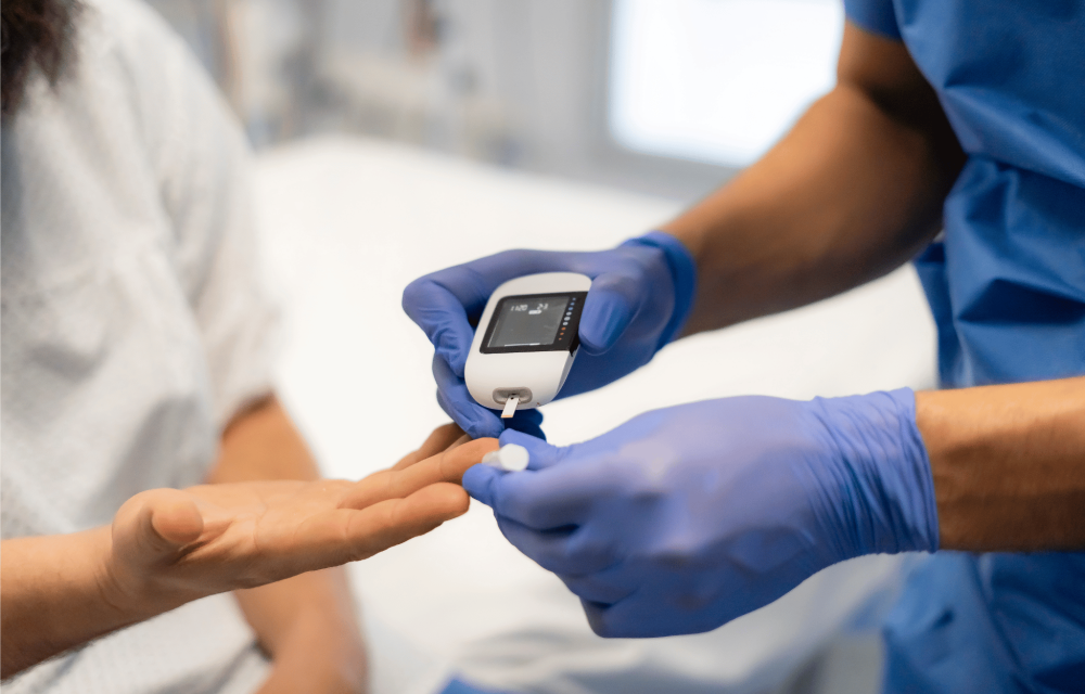 Healthcare worker wearing blue gloves uses a handheld glucose meter to check a patient’s blood sugar from a finger prick.