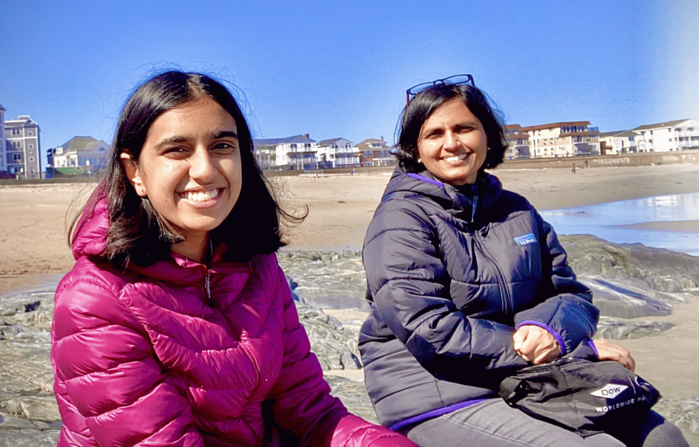 Two people in puffer jackets sit on rocks at a beach with beachfront homes and a clear blue sky in the background.