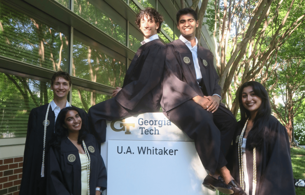 Five graduates wearing academic regalia pose around a Georgia Tech U.A. Whitaker building sign outdoors. Two graduates sit on top of the sign while three stand beside it. A glass building and leafy trees surround the group, with sunlight filtering through the branches. Visible text on the sign reads “Georgia Tech” and “U.A. Whitaker.”