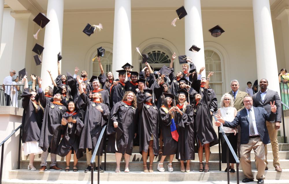 Graduates of the MBID program in Coulter BME are standing on the steps of the Historic Academy of Medicine with their caps and gowns on tossing their caps in the air. The department chair, two faculty, and the program director or standing on the right side of the steps. 