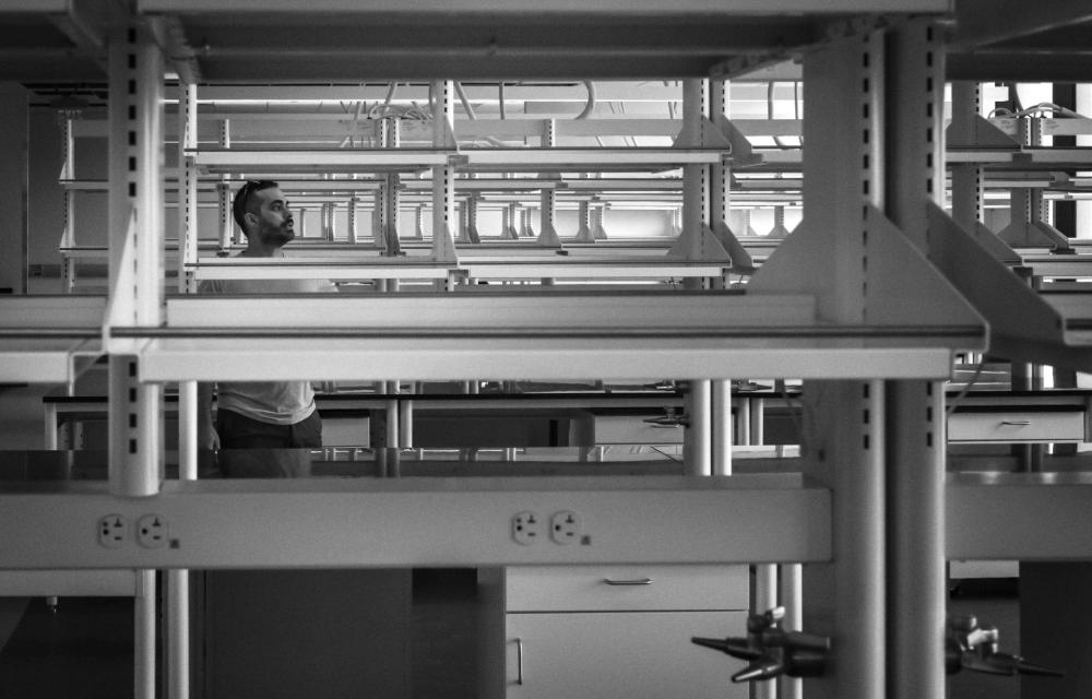 A person stands between rows of laboratory benches and shelving in a black-and-white research lab photograph.