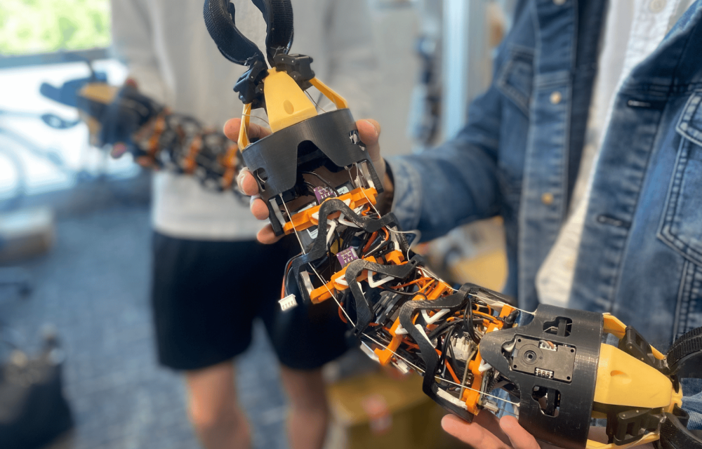 Close-up of a modular, snake-like robotic device with exposed wiring, sensors, and claw-like grippers at both ends being held by two people in a laboratory setting. The flexible robotic structure features black and orange components connected by segmented joints.